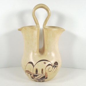 May include: A cream-colored, double-spouted pottery vase with a handle. The vase features a hand-painted design of a bird with geometric patterns in brown and black. The vase is approximately 10 inches tall.