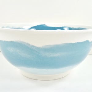 May include: A white ceramic bowl with a wide, light blue painted band around the middle. The bowl has a simple, modern design, perfect for serving salads or displaying fruit. The bowl is approximately 25 cm in diameter.