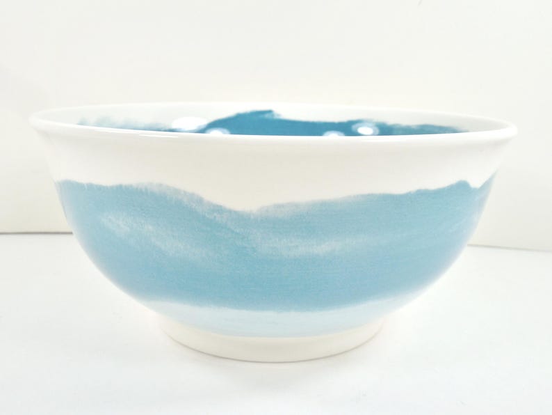 May include: A white ceramic bowl with a wide, light blue painted band around the middle. The bowl has a simple, modern design, perfect for serving salads or displaying fruit. The bowl is approximately 25 cm in diameter.