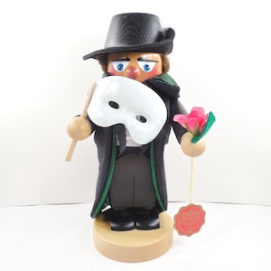May include: A wooden nutcracker figurine of a man in a black top hat and coat, holding a white mask and a pink flower. The figurine has a painted face with blue eyes and red cheeks. The coat has green trim.