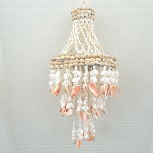 May include: A handcrafted chandelier made of various seashells. The chandelier features a circular design with multiple strands of shells hanging down. The shells are primarily white and beige, with some coral-colored accents. The fixture is suspended from a white cord.