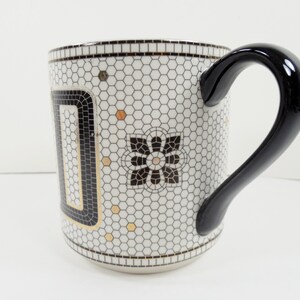 May include: A ceramic mug with a black handle and a white background. The mug features a mosaic-like pattern of small hexagons in white and black, with gold accents and a black floral design. The handle is glossy black.