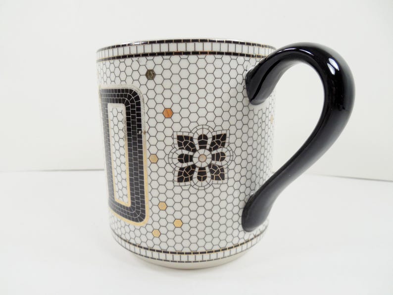 May include: A ceramic mug with a black handle and a white background. The mug features a mosaic-like pattern of small hexagons in white and black, with gold accents and a black floral design. The handle is glossy black.