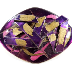 May include: A vintage brooch with a geometric design. The brooch features a deep purple base with gold and pink accents. Overlaid are purple and gold rectangular shapes, creating a layered effect. The brooch has a unique, artistic appearance.