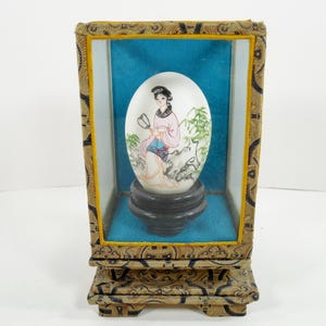 May include: An egg-shaped porcelain figurine, hand-painted with an Asian woman holding a fan, displayed in a decorative wooden frame with a blue background. The frame is ornate with gold accents and rests on a matching base.