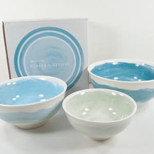 May include: A set of three ceramic bowls with a watercolour design in shades of blue and white. The bowls have a smooth, glossy finish. The box behind the bowls has the text "PEPPER + VETIVER".