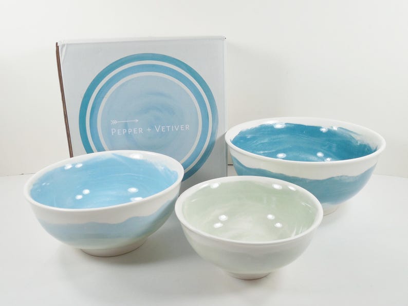 May include: A set of three ceramic bowls with a watercolour design in shades of blue and white. The bowls have a smooth, glossy finish. The box behind the bowls has the text "PEPPER + VETIVER".