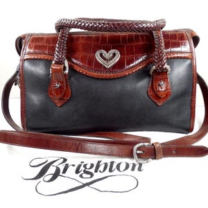 May include: A two-tone handbag featuring a black leather body and a brown crocodile-embossed leather top flap. It includes a braided brown leather handle, a detachable shoulder strap, and a silver-tone heart-shaped emblem. The word Brighton is printed below.