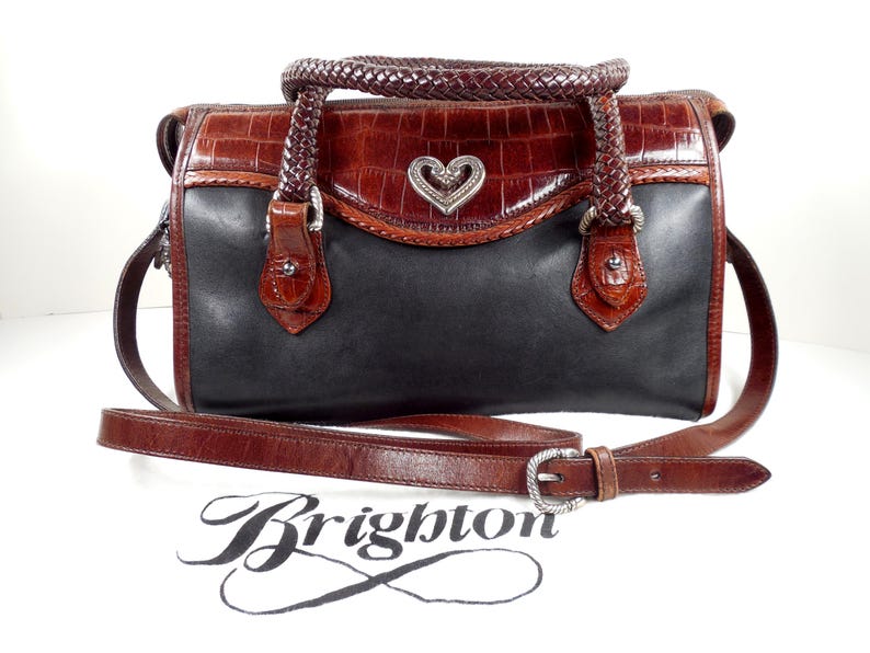 May include: A two-tone handbag featuring a black leather body and a brown crocodile-embossed leather top flap. It includes a braided brown leather handle, a detachable shoulder strap, and a silver-tone heart-shaped emblem. The word Brighton is printed below.