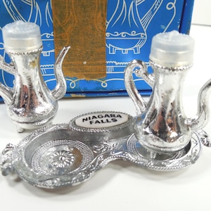May include: A silver-colored salt and pepper shaker set shaped like small teapots. The set includes two shakers with clear plastic tops and a decorative tray with the words "Niagara Falls". The set is displayed in front of a blue box.