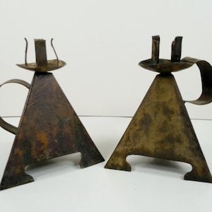 May include: Two vintage metal candle holders. Each has a triangular base with a curved handle and a small dish to hold the candle. The candle holders are made of a dark, aged metal with a patina finish. The overall design is simple and geometric.