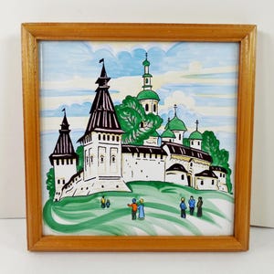 May include: A square ceramic tile featuring a hand-painted scene of a white fortress with dark brown roofs and green domes, set against a blue sky with clouds. The tile is framed in a light brown wooden border and depicts small figures.