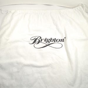 May include: A white fabric drawstring dust bag featuring the black text Brighton printed in a stylized script font in the center. The bag has a gathered top edge with a thin white cord closure visible on the left and right sides of the opening.