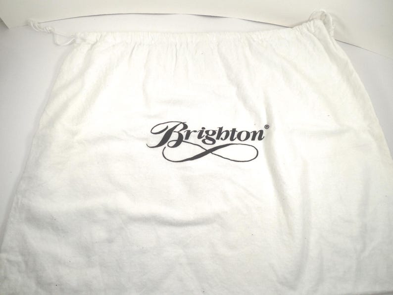 May include: A white fabric drawstring dust bag featuring the black text Brighton printed in a stylized script font in the center. The bag has a gathered top edge with a thin white cord closure visible on the left and right sides of the opening.