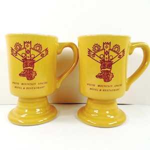 May include: Two yellow ceramic pedestal mugs featuring a maroon graphic design of a crown-like symbol above the text White Mountain Apache Motel & Restaurant. Each mug has a sturdy handle and a wide, circular base for stability on flat surfaces.