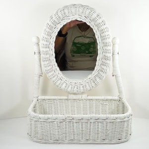 May include: White wicker vanity mirror with an oval frame and a rectangular basket base. The mirror is supported by two vertical posts. The frame, posts, and basket are all made of woven wicker.