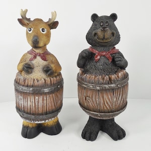 May include: Two decorative figurines, a deer and a bear, are standing in wooden barrels. Both figurines are wearing red bandanas around their necks.