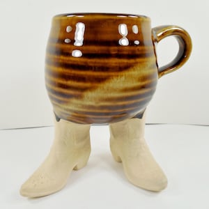 May include: A unique ceramic mug with a brown and tan striped design, featuring a handle and two beige cowboy boot legs as a base. The mug has a glossy finish and is designed to stand upright.