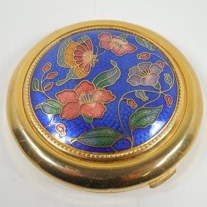 May include: A gold compact with a blue enamel lid featuring a floral design with butterflies. The flowers are pink, red, and purple.