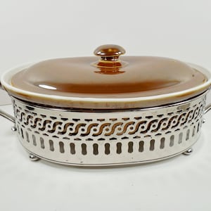 May include: A brown ceramic casserole dish with a silver metal stand. The dish has a lid and two handles. The stand has a decorative design.