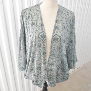 May include: A light blue open-front cardigan featuring a dark blue damask pattern. The garment has a relaxed fit with three-quarter length sleeves and a draped silhouette, showcasing a textured velvet-like finish throughout the fabric.