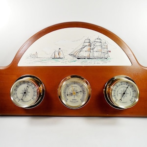May include: A wooden weather station featuring a thermometer, barometer, and hygrometer with brass-toned circular frames. The upper section displays a white ceramic panel with a detailed illustration of two sailing ships on the ocean.