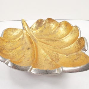 May include: A decorative leaf-shaped bowl with a metallic gold interior and a silver-colored rim. The bowl has a textured surface and is designed to resemble a leaf. The bowl is likely used for decorative purposes.