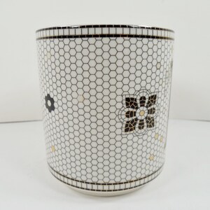 May include: White ceramic tumbler with a black and white honeycomb pattern. The tumbler features black and gold floral designs. The rim and base have a black mosaic tile design.