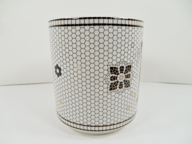 May include: White ceramic tumbler with a black and white honeycomb pattern. The tumbler features black and gold floral designs. The rim and base have a black mosaic tile design.
