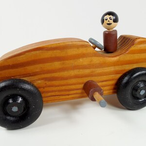 May include: A wooden toy car with black wheels and a painted wooden figure in the driver's seat. The car has a brown painted steering wheel and a gray painted exhaust pipe.