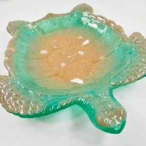 May include: A decorative, turtle-shaped dish with a textured surface. The dish features a vibrant color palette, with a turquoise rim and a sandy-colored interior. The dish has a glossy finish and is designed to resemble a turtle.