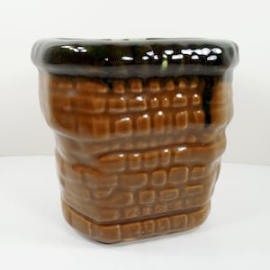 May include: A brown ceramic planter with a textured brick pattern. The top rim is dark brown and glossy. The planter has a square base and a slightly wider top, suitable for indoor or outdoor use.