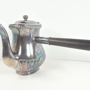 May include: A vintage silver-plated coffee pot with a dark wooden handle. The pot has a decorative design around the base and lid, and a curved spout. The handle is attached horizontally to the side of the pot.