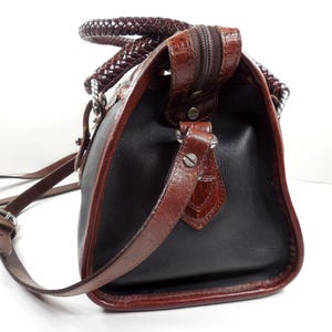May include: A black handbag with brown trim and a textured crocodile-embossed finish on the edges. The bag features a braided brown handle, a top zipper closure, and a thin adjustable brown shoulder strap attached with silver-toned metal hardware.