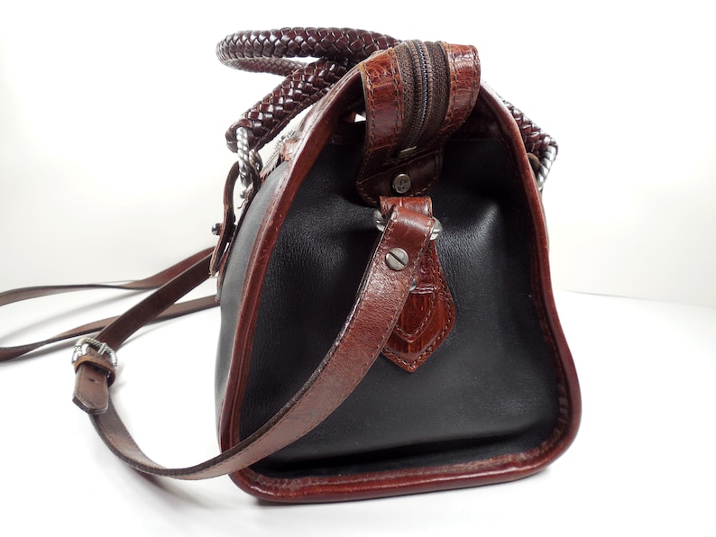 May include: A black handbag with brown trim and a textured crocodile-embossed finish on the edges. The bag features a braided brown handle, a top zipper closure, and a thin adjustable brown shoulder strap attached with silver-toned metal hardware.