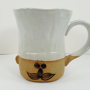 May include: A ceramic mug with a chef-themed design. The top portion is white, and the bottom is tan with a cartoon face. The mug has a curved handle and is suitable for hot or cold beverages.