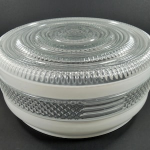 May include: A clear glass light fixture with a white base. The top is a clear, circular design with concentric rings. The middle section has a textured pattern. The fixture is round and sits on a dark surface.