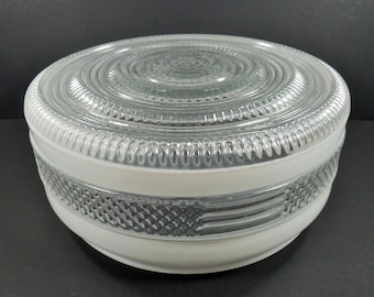 May include: A clear glass light fixture with a white base. The top is a clear, circular design with concentric rings. The middle section has a textured pattern. The fixture is round and sits on a dark surface.