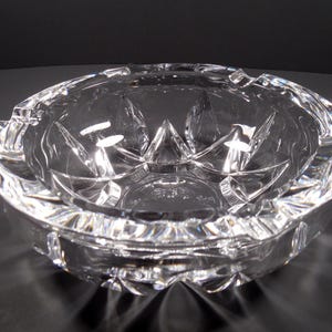 May include: A clear, round crystal ashtray with a starburst pattern on the base and sides. The ashtray has a scalloped rim with notches for resting cigarettes. The glass reflects light, creating a sparkling effect.