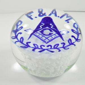 May include: A clear glass paperweight with a blue and white design. The design features a compass and square, a symbol of Freemasonry, surrounded by a wreath. The text "F. & A. M." is inscribed around the design.