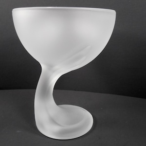 May include: A frosted glass goblet with a unique, curved stem and a wide bowl. The stem gracefully swirls into a circular base, creating an elegant and artistic design. The glass has a smooth, matte finish.