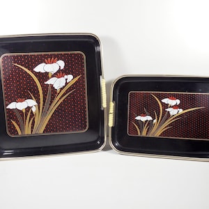 May include: Two black serving trays with gold trim and yellow handles. Each tray features a floral design with white flowers, gold stems, and a red dotted background. The trays are rectangular, one square and one smaller.
