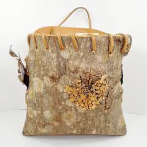 May include: A rustic, handmade wall basket crafted from a section of tree trunk. The bark exterior is a mix of brown and tan, with visible texture and natural imperfections. The top edge is lined with light-colored wood and leather stitching, and a leather handle is attached.