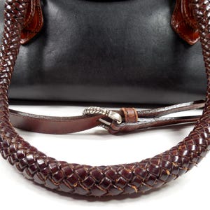 May include: A black handbag featuring dark brown braided leather handles and a matching brown leather shoulder strap with a silver-toned metal buckle. The bag has a smooth black body with brown leather accents at the handle attachment points.