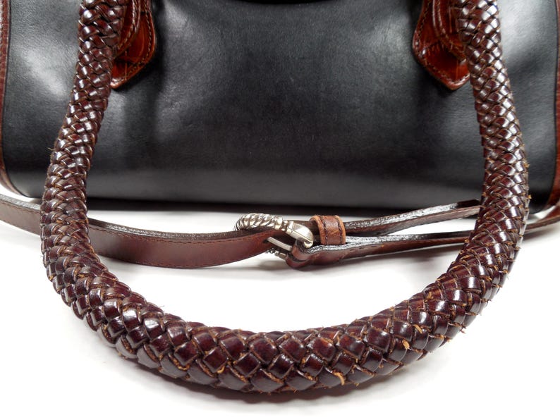 May include: A black handbag featuring dark brown braided leather handles and a matching brown leather shoulder strap with a silver-toned metal buckle. The bag has a smooth black body with brown leather accents at the handle attachment points.