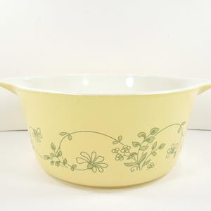 May include: A vintage yellow Pyrex mixing bowl with a white interior and two handles. The exterior features a floral design in shades of green. This classic kitchenware is perfect for baking and food preparation.