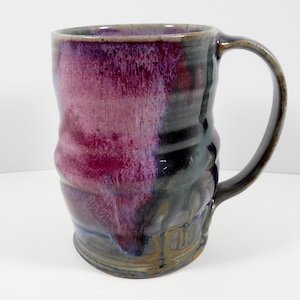 May include: A handmade ceramic mug with a dark purple and black glaze. The mug has a unique, slightly uneven shape and a handle.