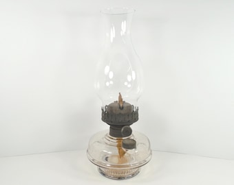 May include: A clear glass oil lamp with a tall, curved chimney. The lamp has a round base and a metal burner assembly. The lamp is designed to hold oil and has a wick for lighting. The lamp is a vintage style.
