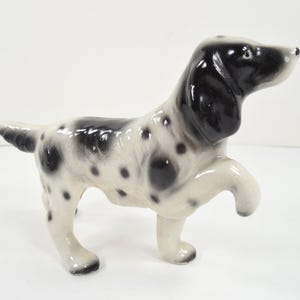 May include: A ceramic figurine of a dog with black and white markings. The dog is posed with one paw raised, giving the appearance of walking. The dog's ears and head are black, and its body is white with black spots.