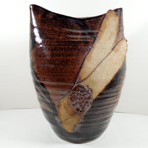 May include: A handcrafted ceramic vase with a unique design. The vase features a dark brown and black color scheme with a textured surface. A tan-colored, leaf-like accent is attached to the side. The vase is approximately 12 inches tall.